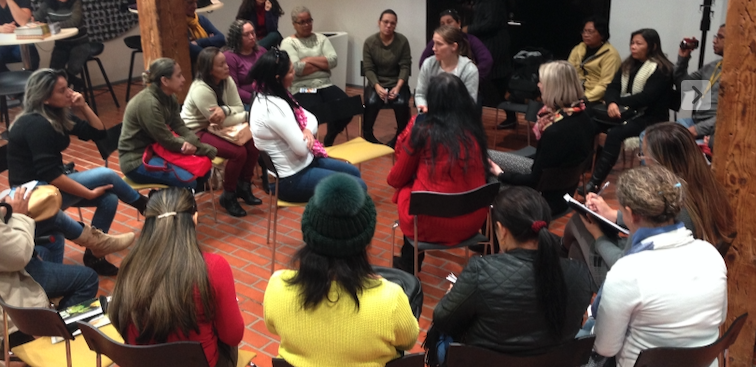 Diverse group of adults seated in a circle, engaged in a discussion during a community meeting in a brick-floored room. ] , (fix)